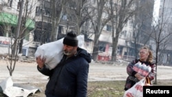 在被俄军围攻的南部港城马里乌波尔,当地居民肩扛手提,携带着食物走过被战火破坏的一座居民楼。(2022年3月31日)