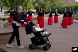资料照:一名老年妇女推着一个婴儿走过北京一所公园里跳广场舞的妇女。(2021年10月14日)