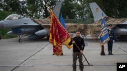 资料照片:在一处未披露的地点,乌克兰总统泽连斯基把一支部队的军旗授予该部队,他的身后是乌克兰空军的F-16战斗机。(2024年8月4日)