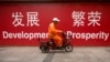 FILE - A maintenance worker rides a scooter past banners reading "Development" and "Prosperity" in English and Chinese on a street in central Beijing, July 15, 2015.